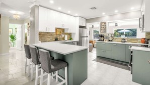 Une cuisine moderne avec des meubles blancs, un îlot central doté de tabourets, un réfrigérateur et une fenêtre donnant sur un espace verdoyant.