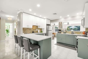 Une cuisine moderne avec des meubles blancs, un îlot central doté de tabourets, un réfrigérateur et une fenêtre donnant sur un espace verdoyant.