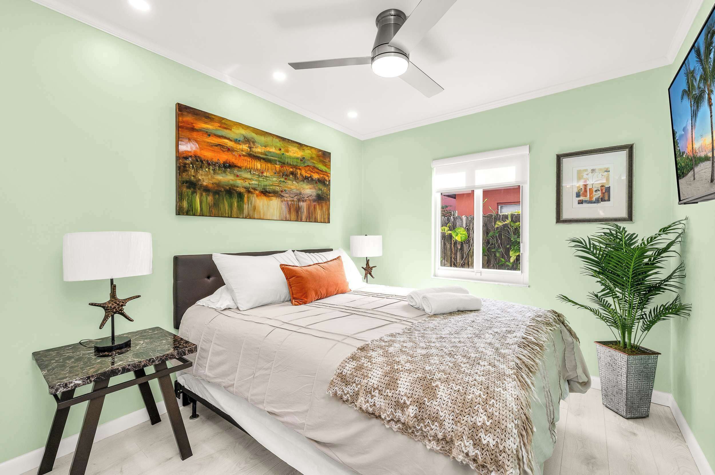 A bedroom with a bed, two bedside tables, a ceiling fan, a window, and a potted plant.