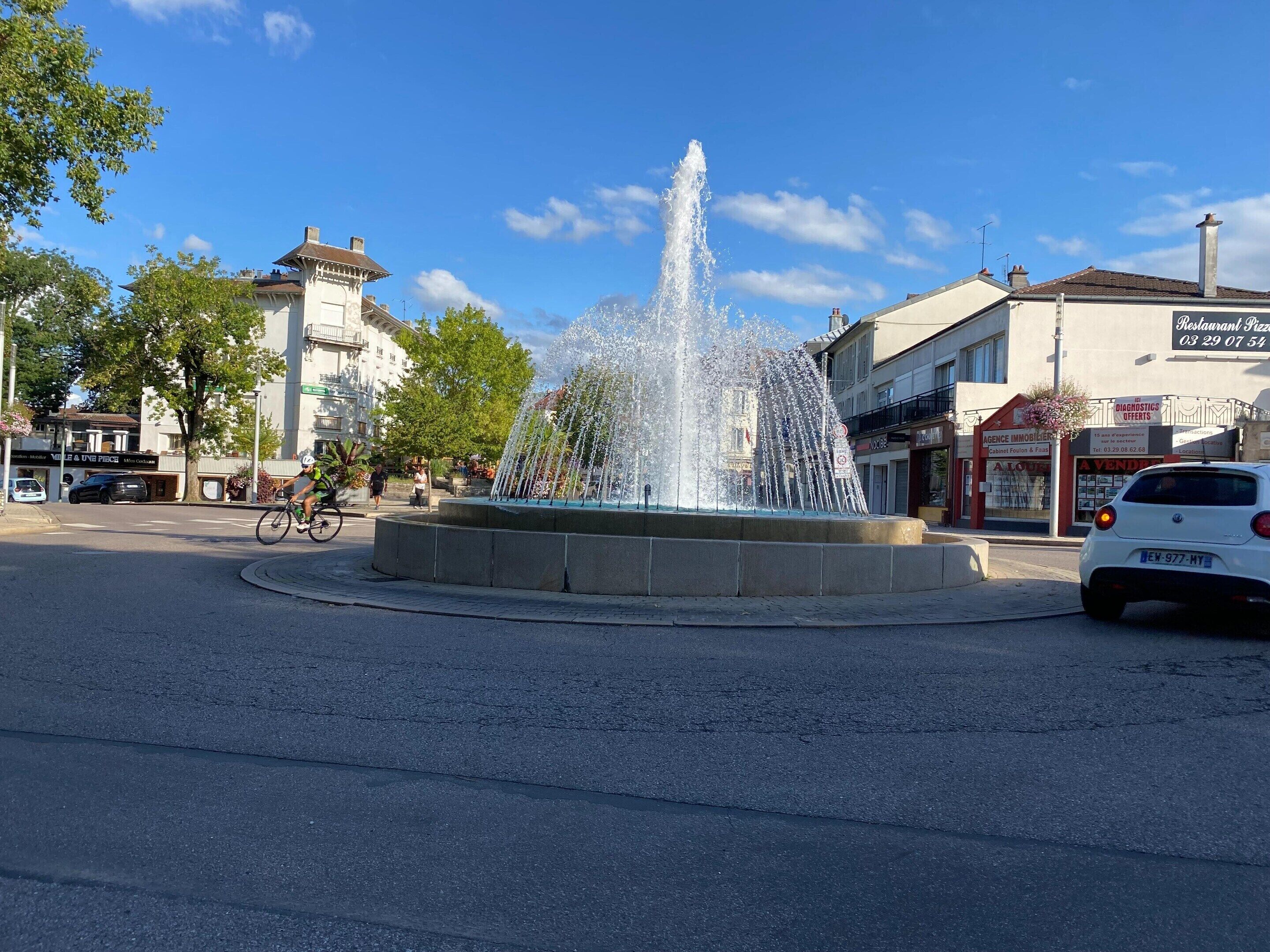 Une fontaine publique avec des jets d’eau, entourée d’une zone pavée, avec des bâtiments et une voiture en arrière-plan.