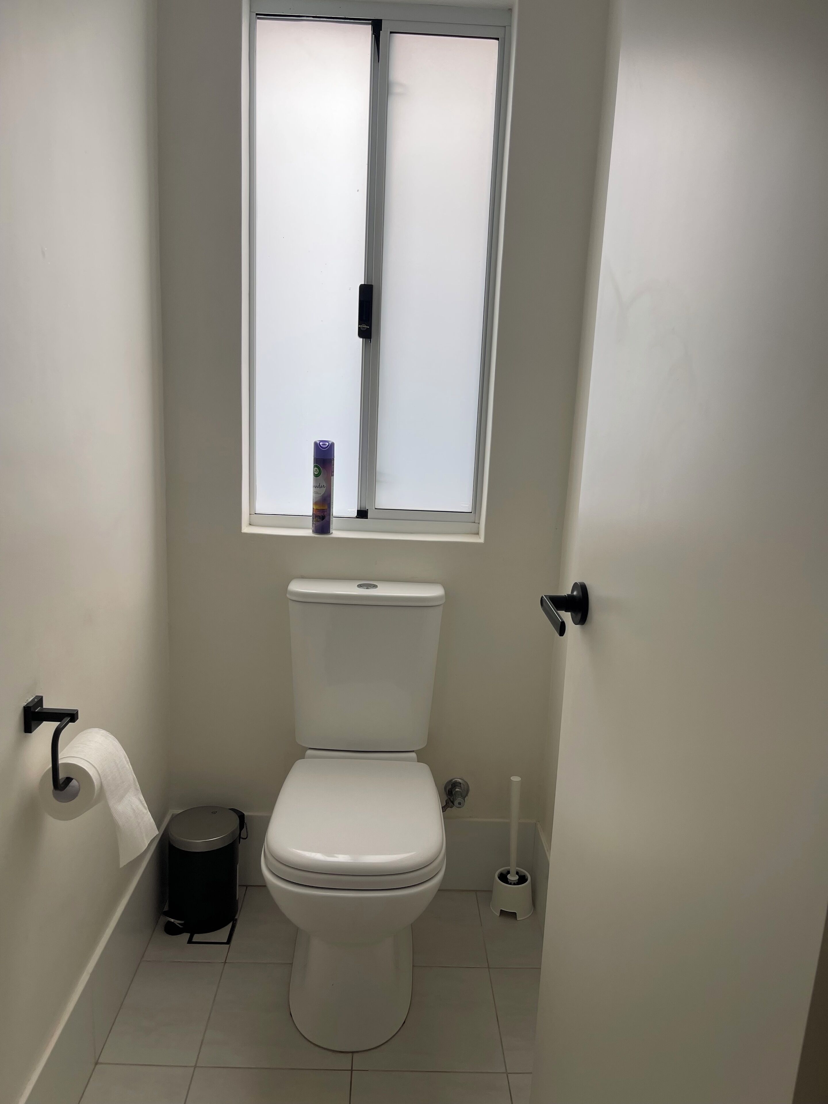 A small, clean bathroom with a toilet, a trash bin, a toilet paper holder, and a window above the toilet.