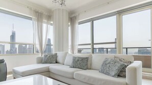 A modern living room with a sectional sofa, a coffee table, and a view of a city skyline through large windows. - StayShort - Dubai Marina sleeps 4 with Marina Views! (Dubai Marina)