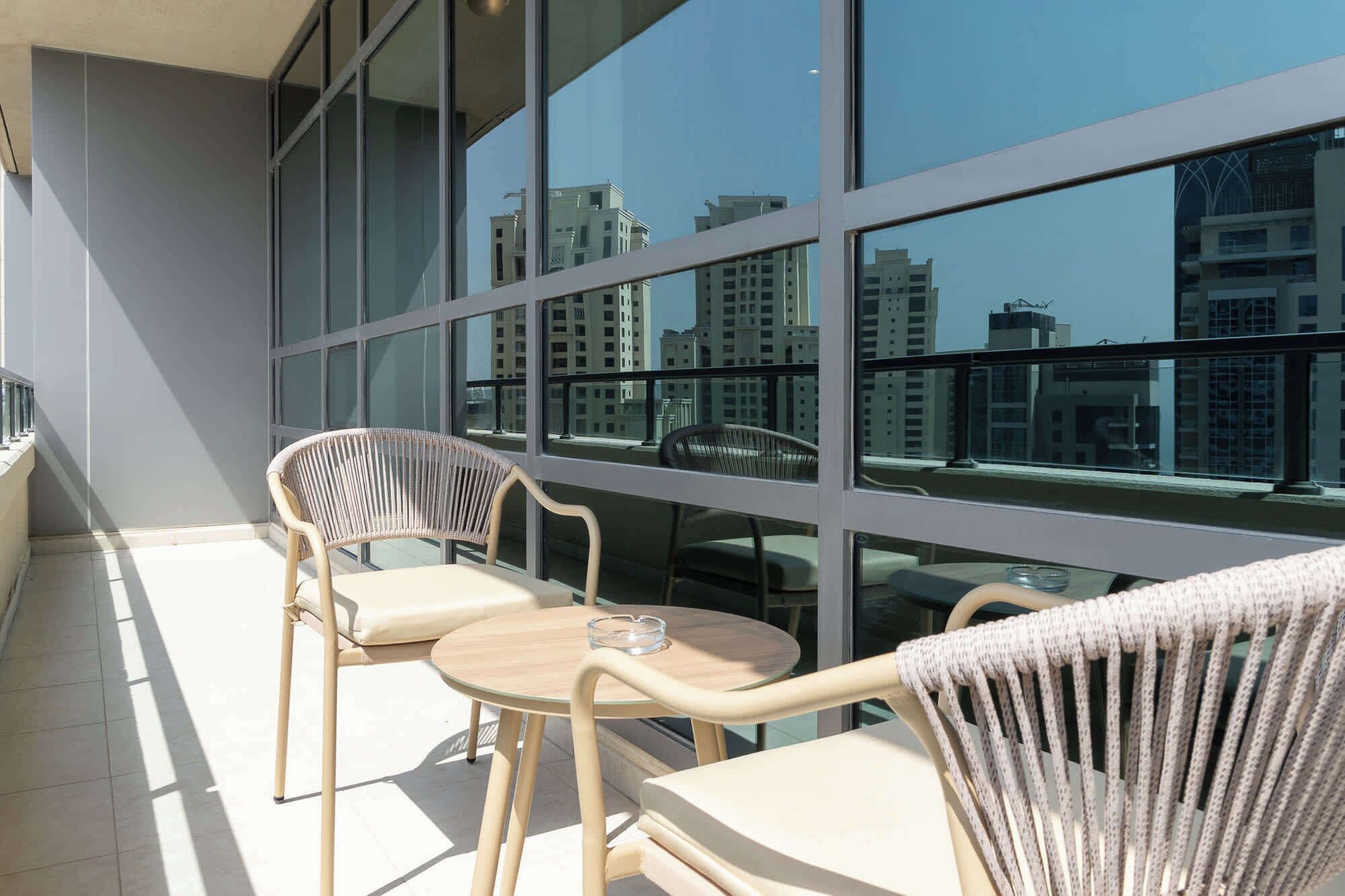A balcony with two wicker chairs and a small table, large windows offering a view of high-rise buildings.