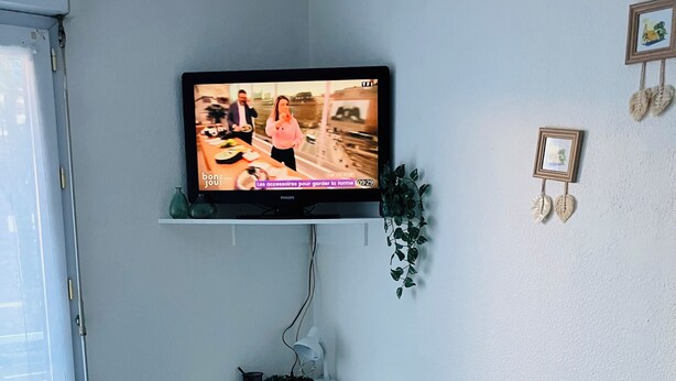 Een moderne kamer met een flatscreen televisie aan een witte muur, een klein wit krukje, een wit geweven vloerkleed en een plant op een plank.