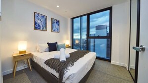 Una camera d'albergo moderna con un letto grande, due comodini con lampade, vista sulla città tramite finestre a tutta parete e due quadri astratti incorniciati.