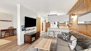 Espaço compacto com televisão, mobiliário de madeira, área de cozinha e um aviso na parede.