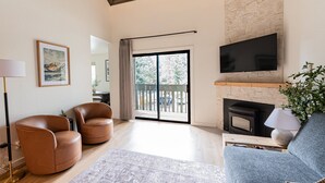 Un vaste salon doté d’une cheminée, d’un téléviseur fixé à un mur en pierre, de fauteuils en cuir et d’une grande porte-fenêtre coulissante donnant sur un balcon.