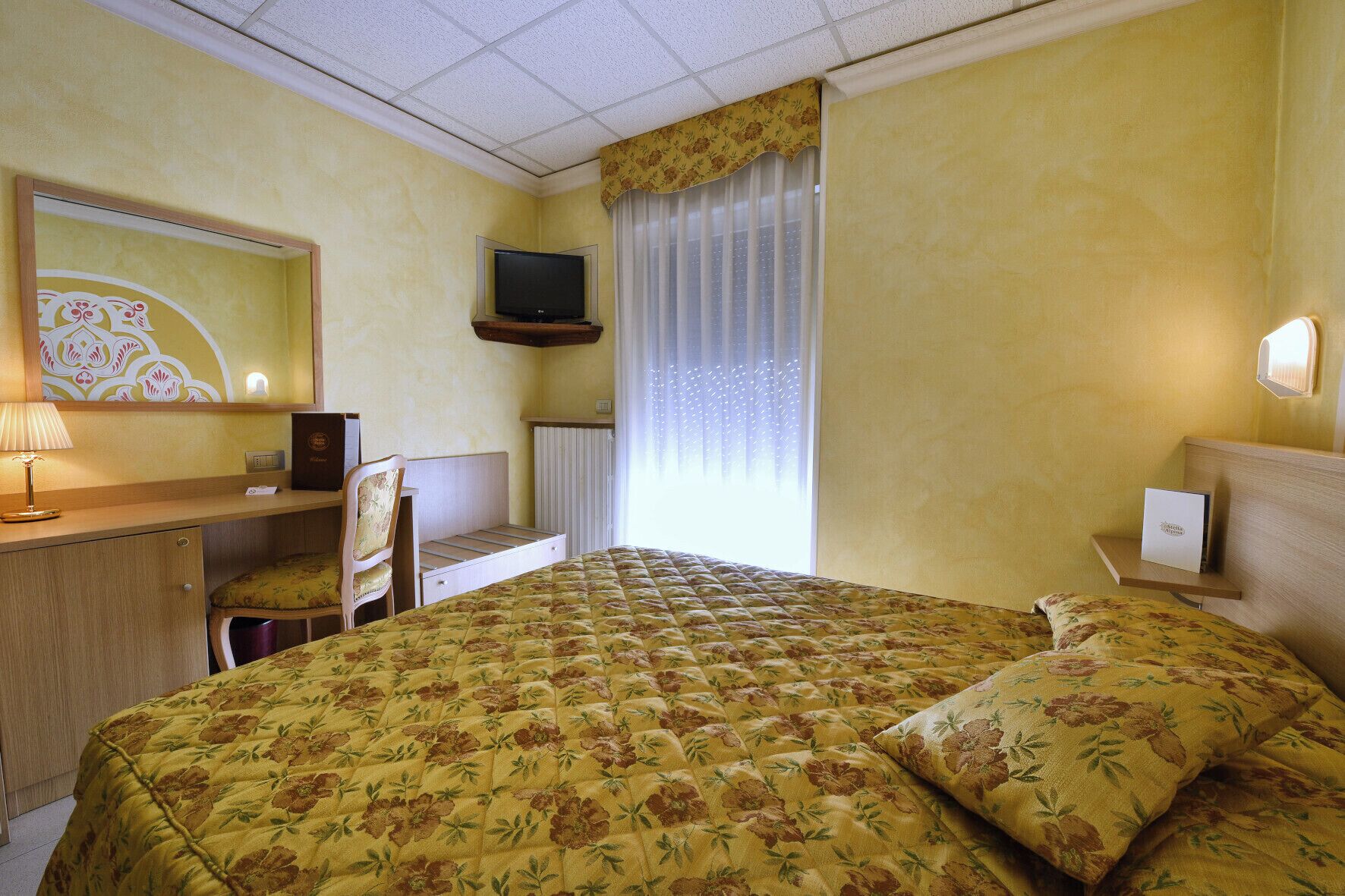 A hotel room with a bed, a desk with a chair, a television, and a window with curtains.