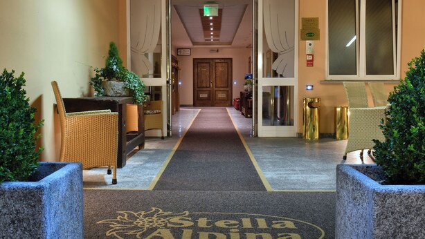 A hotel lobby with a carpeted path leading to a reception area, potted plants, wicker furniture, and a sign reading "Stella Alpina". - Stella Alpina Hotel e Ristorante (Druogno)