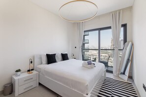 A modern hotel room with a large bed, a bedside table with a lamp, a small plant, and a view of the city through large windows. - Meydan One Sleeps 5 With Great Amenities (Dubai)