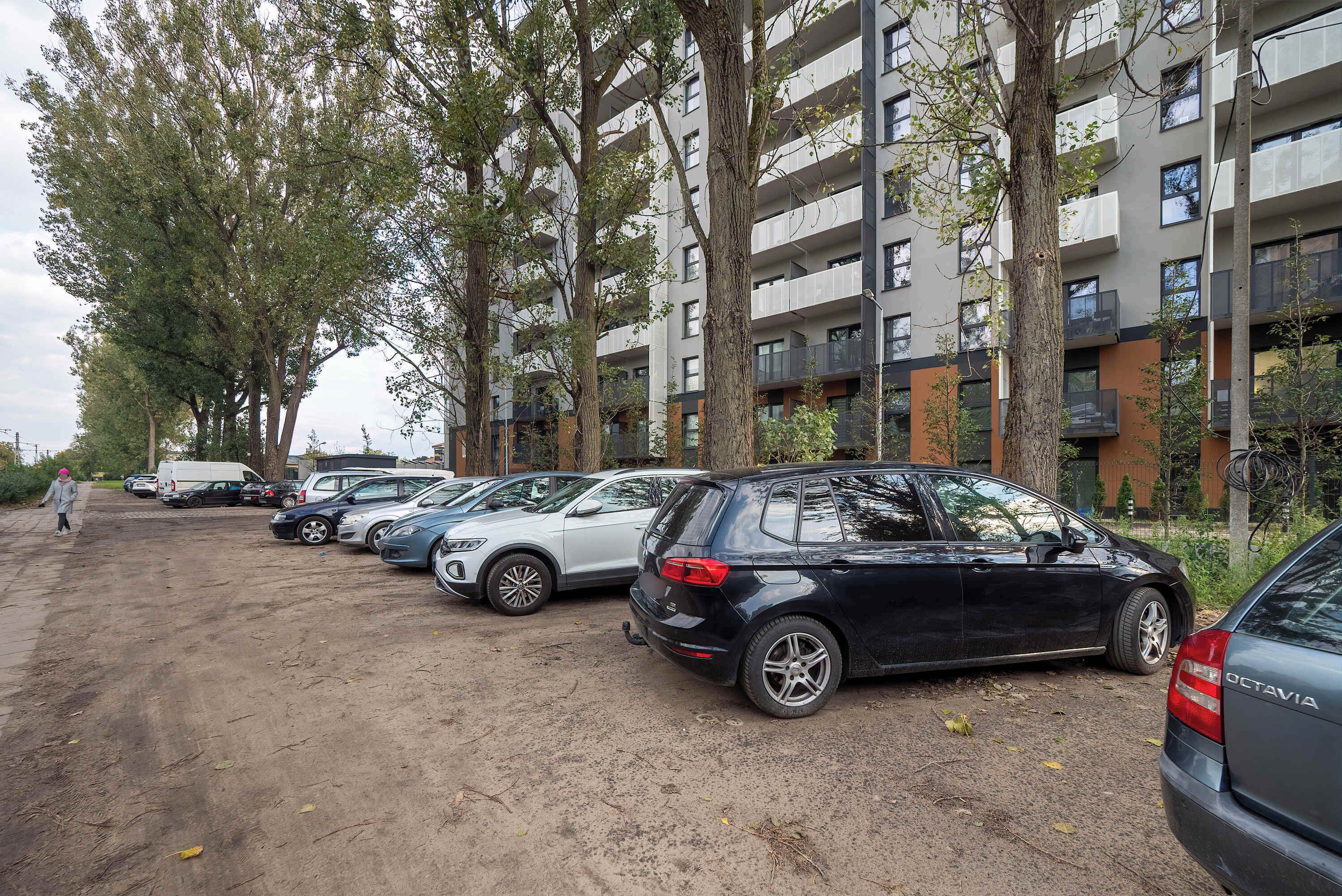 A parking lot with several cars, trees, and a multi-story building with balconies.