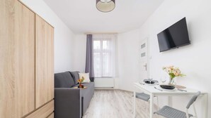 Un espace de vie moderne et compact, avec un canapé gris, une armoire en bois, une petite table à manger avec des chaises, un téléviseur à écran plat et une fenêtre ornée de rideaux légers.