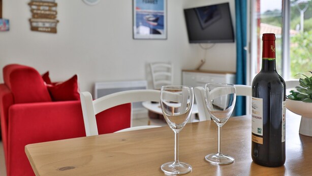 A dining area with a wooden table, two wine glasses, and a wine bottle. There are red chairs and a white chair in the background. - Stylish Villa in Parentis-en-born (Parentis-en-Born)