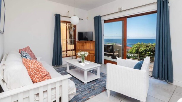 Een moderne woonkamer met een bank, fauteuil en salontafel. Een televisie staat op een houten kast en er is een balkon met uitzicht op zee.