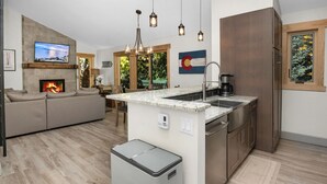 Une cuisine moderne dotée d’un îlot central, d’un four encastré et d’un évier. On y trouve également un coin repas avec une table et des chaises, ainsi qu’un salon avec un canapé et une cheminée.