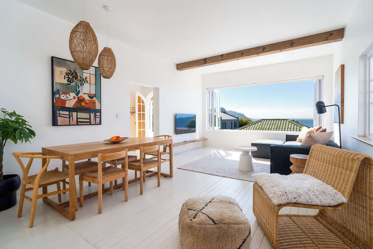 A bright, open living space with a wooden dining table, wicker furniture, and a large window offering a view of the sea.