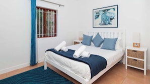 A bedroom with a bed, a window with blue curtains, a wall-mounted painting, and two bedside tables with baskets.