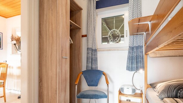 A room with a wooden wardrobe, a chair with a blue cushion, a small table with a radio, and a bed with striped bedding. - Ferienhaus in Busum, 6 Persons (Büsum)
