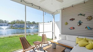 Une terrasse ensoleillée avec vue sur un lac, un canapé blanc, des chaises en bois et des décorations murales représentant des poissons.