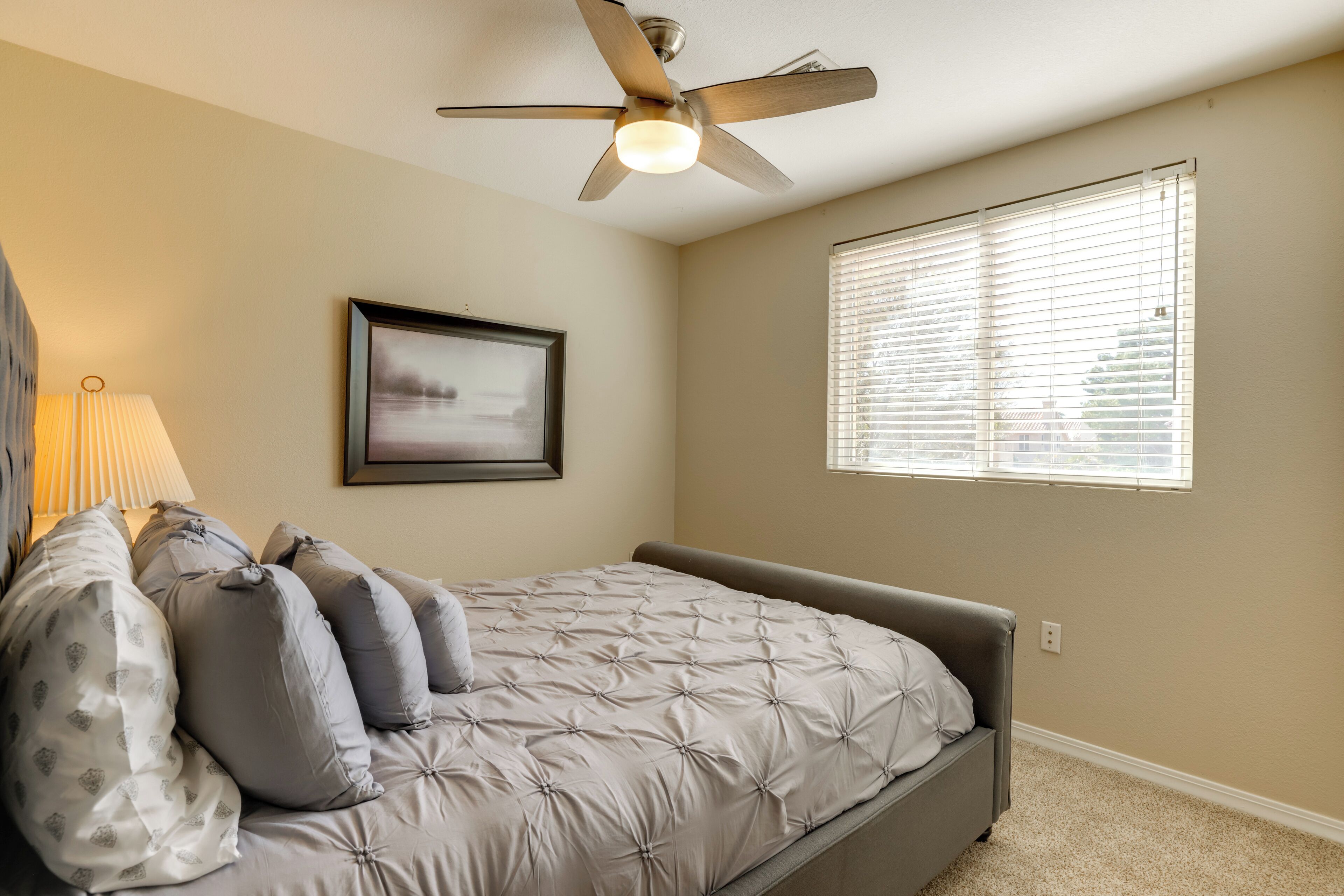 Une chambre à coucher avec un lit, un ventilateur de plafond, une fenêtre avec des stores et un tableau encadré au mur.