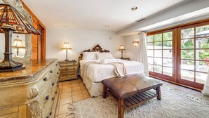 A bedroom with a bed, bedside lamps, a wooden bench, a dresser, and a large window with a view of greenery.