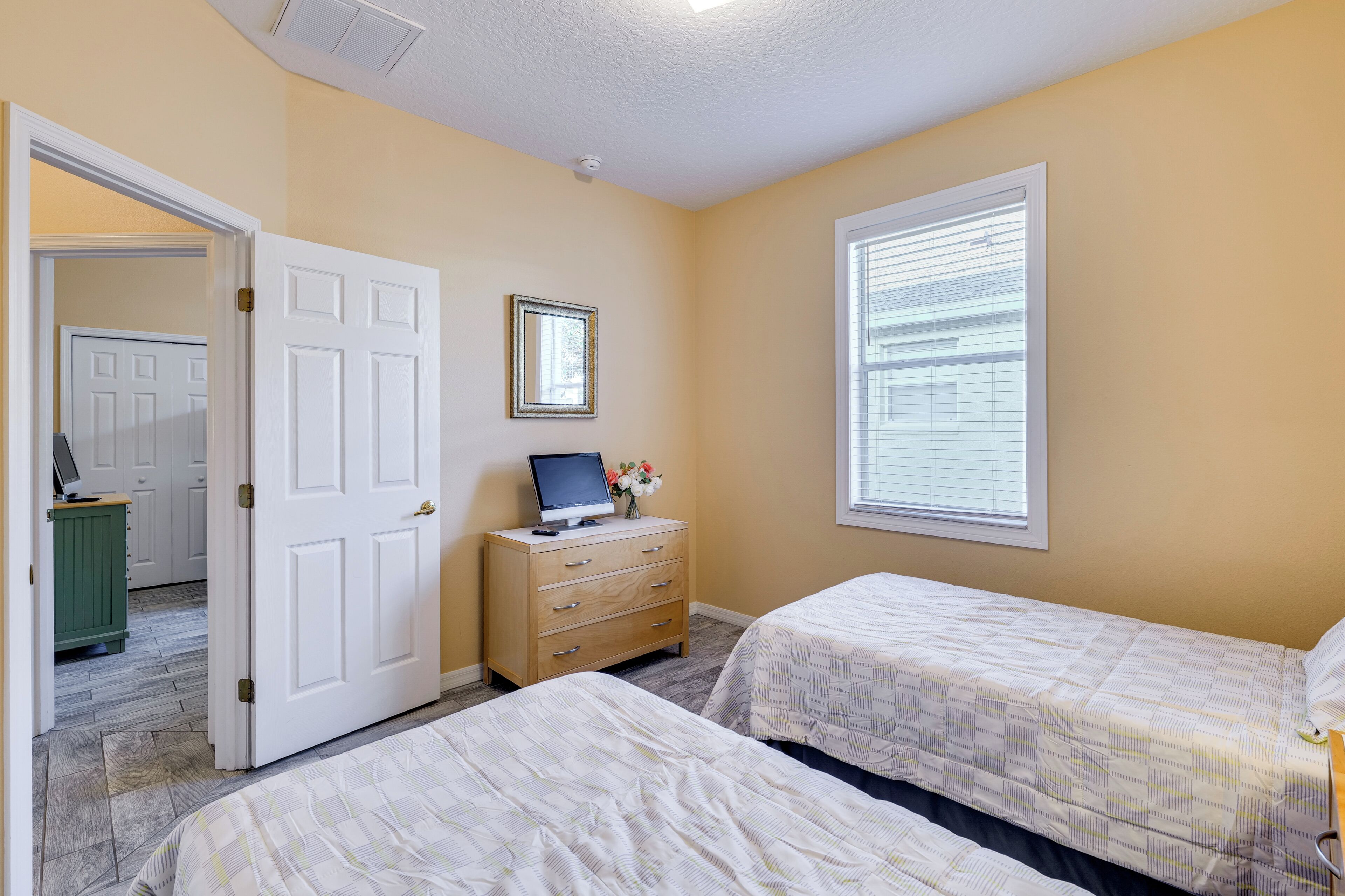 A bedroom with two beds, a dresser, a computer on the dresser, and a window with blinds.