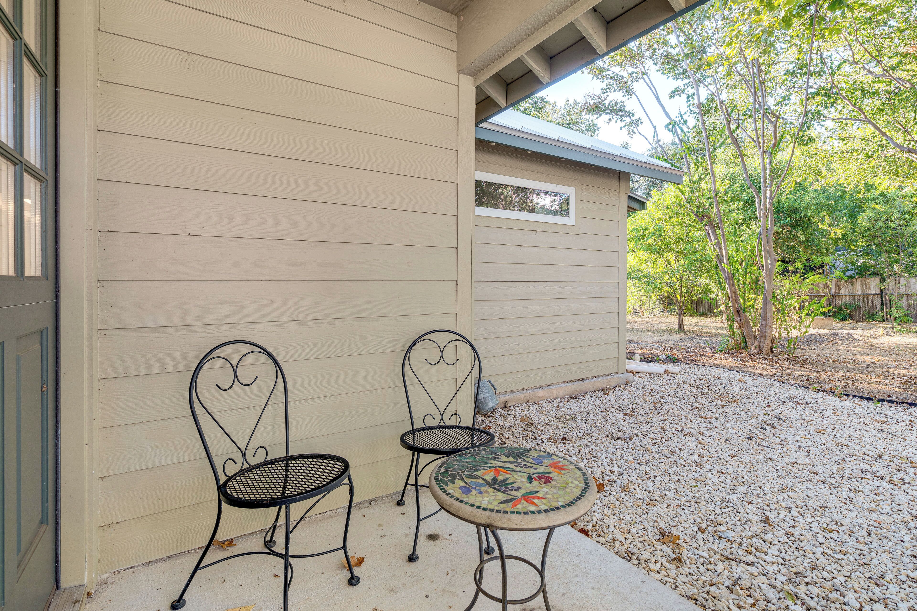 A patio with two metal chairs and a round table with a mosaic top, set against a beige wall with a small window.