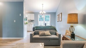 A living room with a sofa, a glass coffee table, and a lamp. There is a kitchen in the background with cabinets and a window. - Home w/ Pet-friendly Yard, 1 Mi to Dtwn Raleigh! (Raleigh)