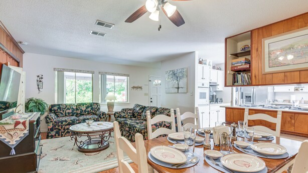 A dining area with a wooden table set for four, surrounded by patterned sofas and a ceiling fan.