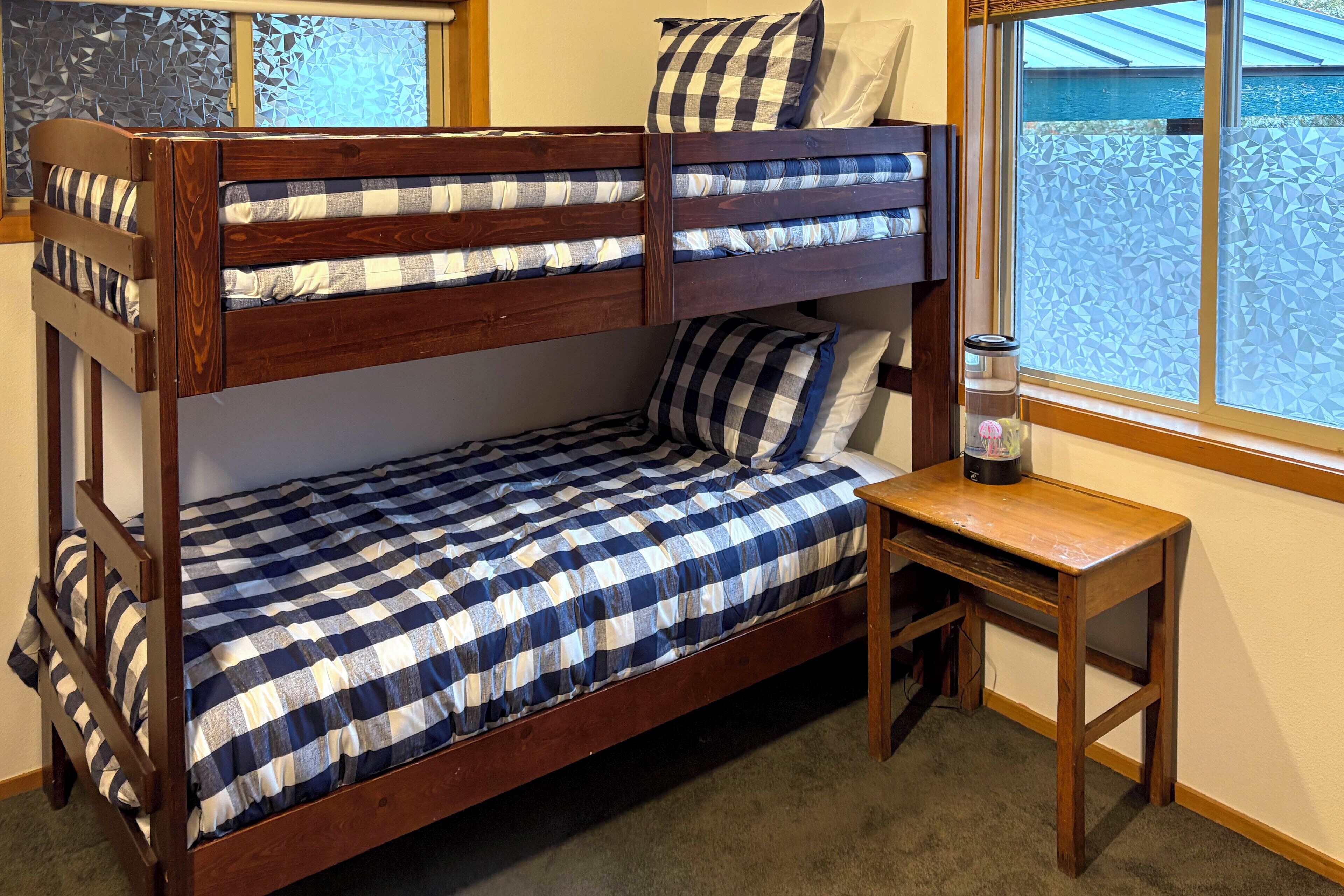 A bunk bed with a checkered bedspread and pillows, a small wooden table, and a window with a view of a blue building.