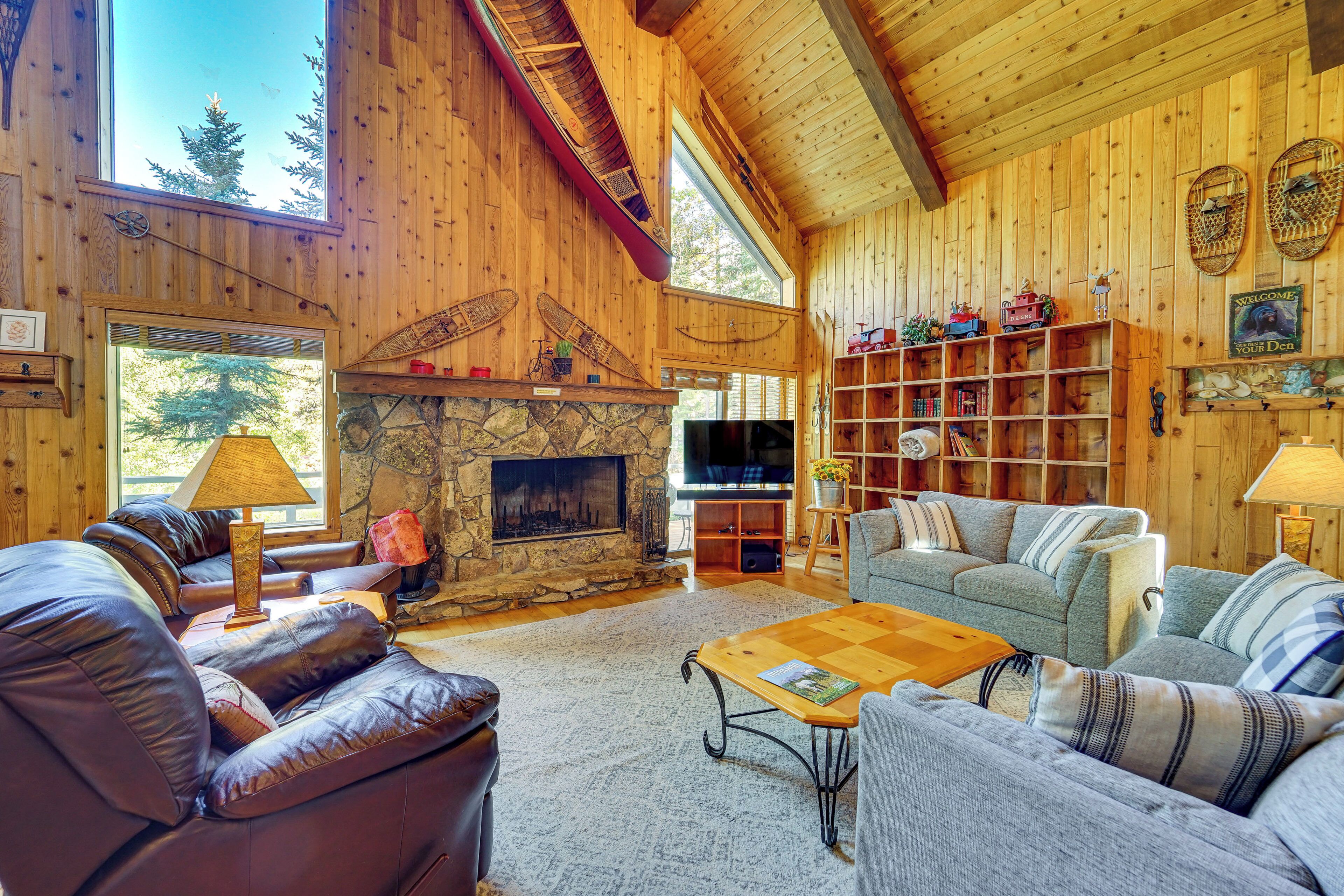 Acogedor interior de cabaña de troncos con chimenea de piedra, sofás de cuero, una mesa de centro de madera y una estantería llena de libros y objetos decorativos.