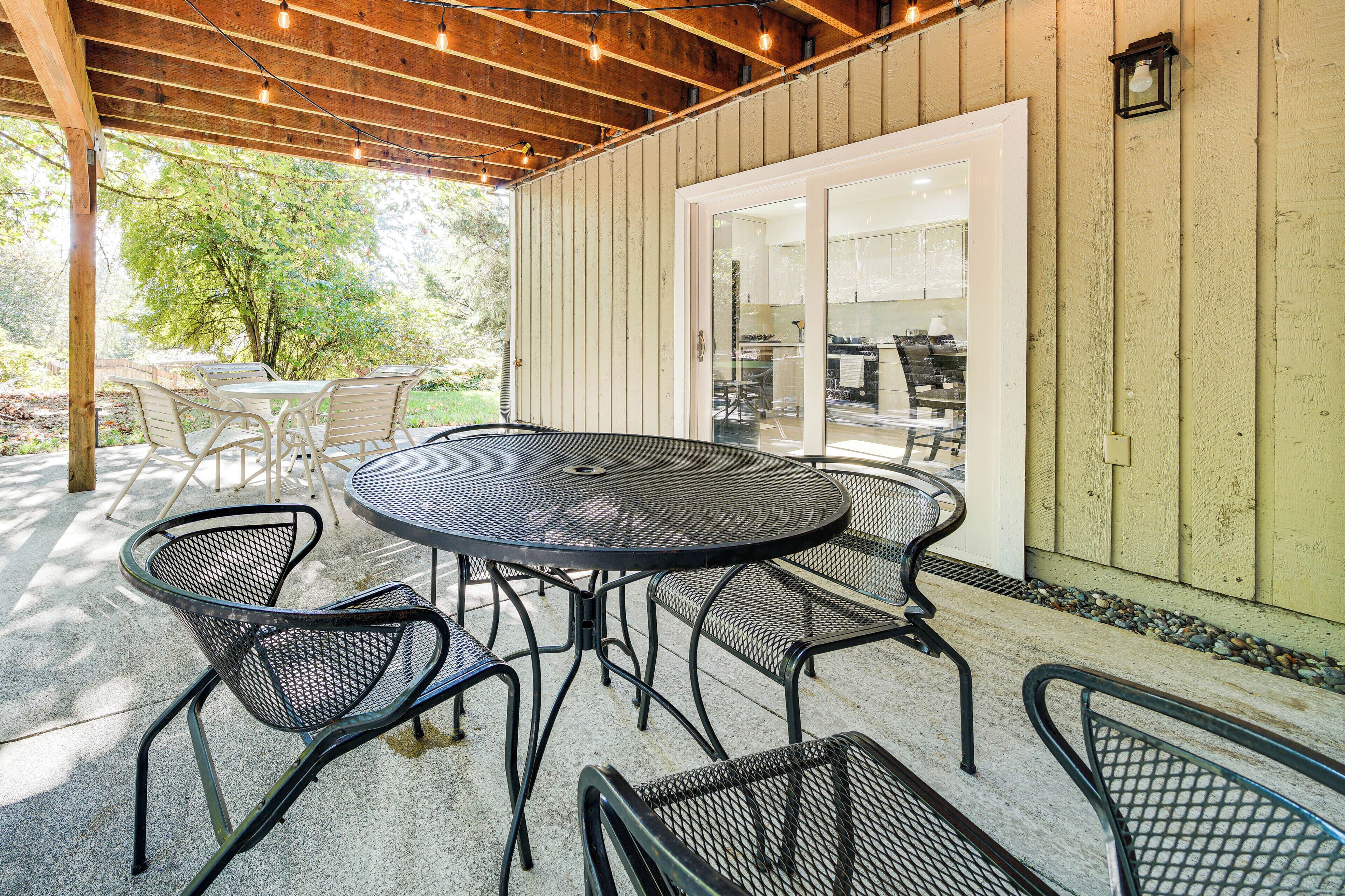 Une terrasse extérieure avec une table ronde et des chaises en métal, des poutres en bois au-dessus et une vue sur l’intérieur à travers une porte vitrée.