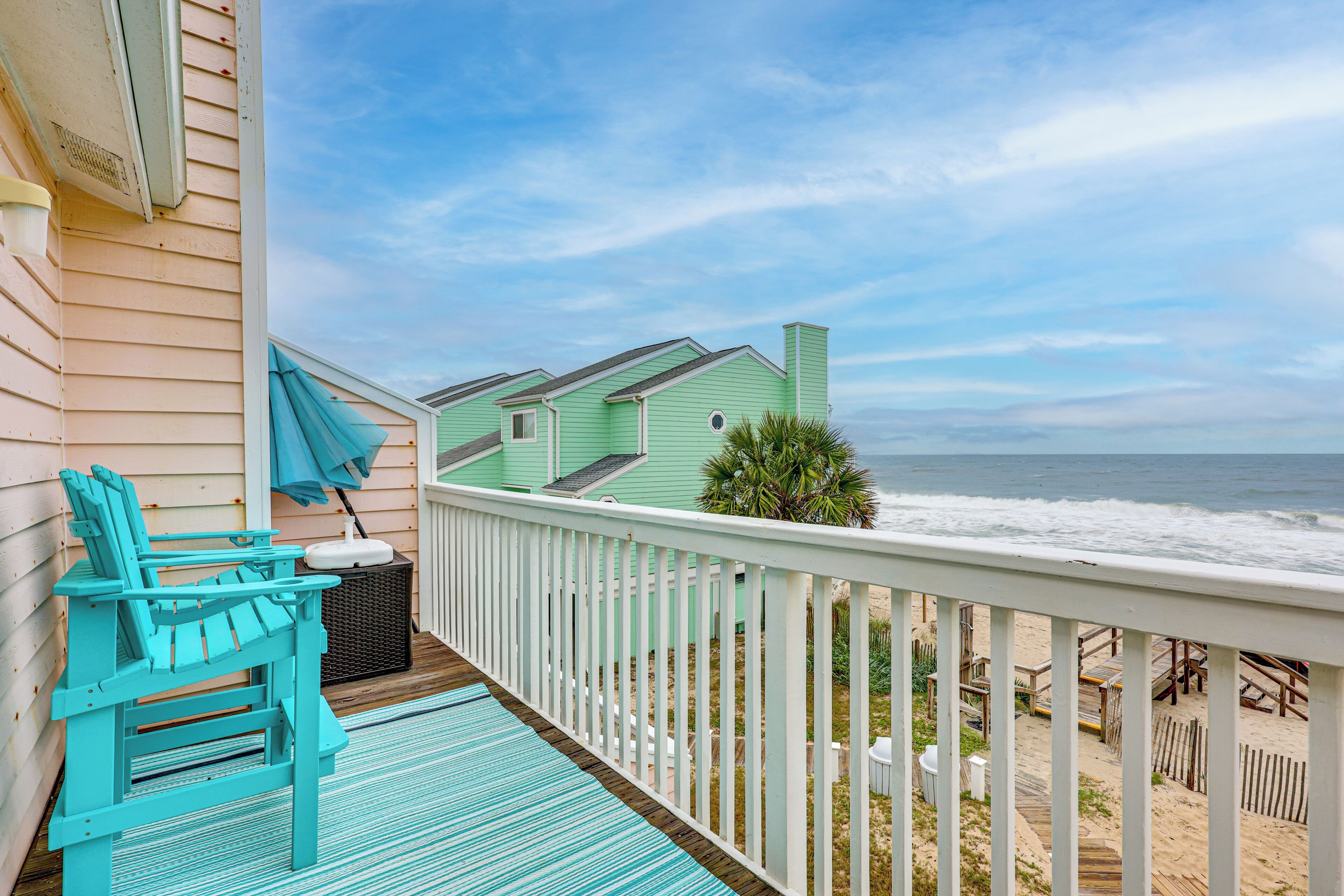 A wooden deck with a turquoise Adirondack chair and a small table, overlooking a beach with a green house and the ocean.