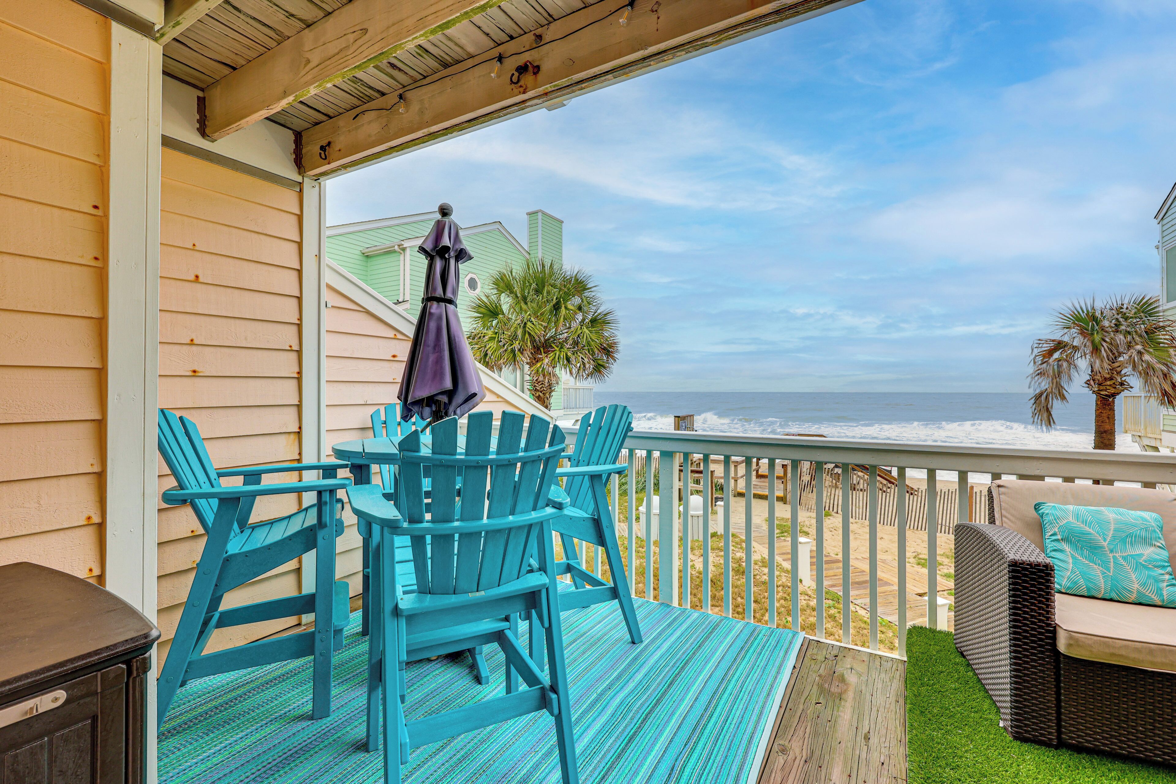 A wooden deck with turquoise chairs and a table, overlooking a beach with palm trees and a view of the ocean.