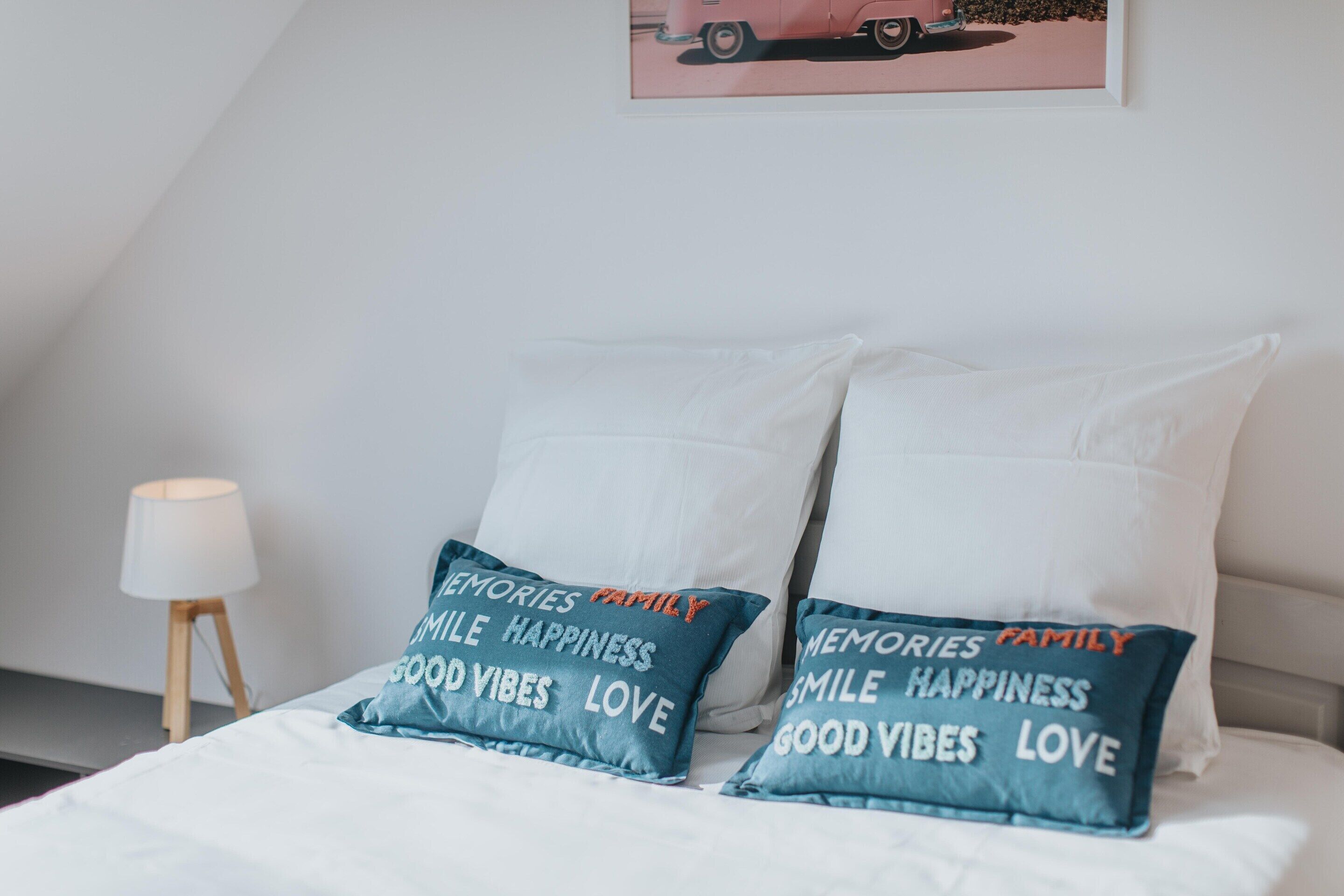 Una cama con ropa de cama blanca y dos almohadas color turquesa con texto, una lámpara de mesa pequeña y un cuadro en la pared.