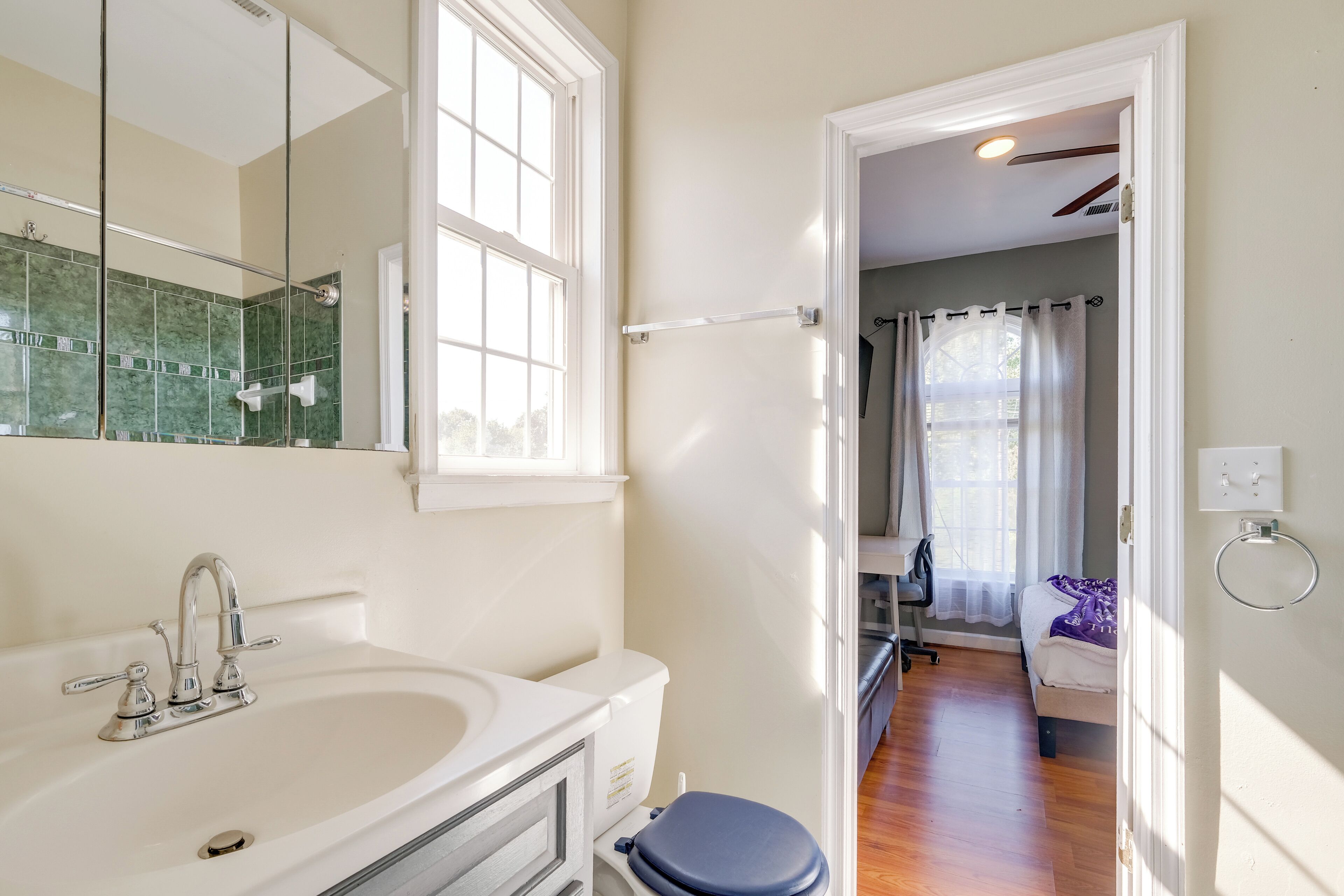 A bathroom with a white sink, a mirror, and a window. There is a door leading to a bedroom with a bed and a chair.
