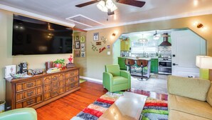 Un salon avec un ventilateur de plafond, un téléviseur, un fauteuil vert et un tapis coloré.
