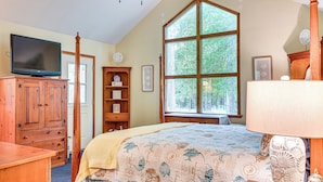 A bedroom with a large window, wooden furniture, a bed with a patterned bedspread, a television on a wooden cabinet, and a bedside table with a lamp. - Forested Family Hideaway Near Lake Sinclair! (Eatonton)