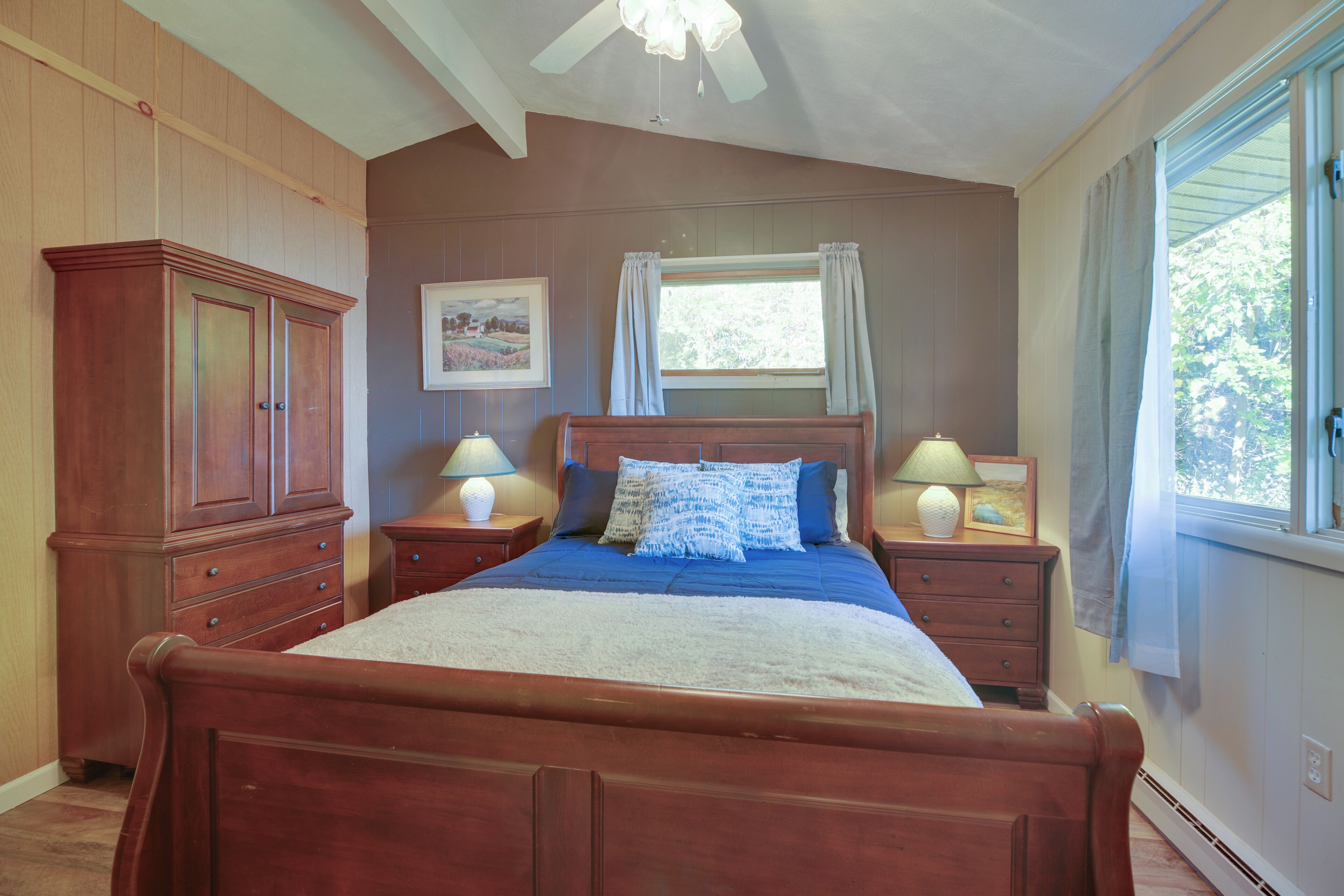 A bedroom with a wooden bed, two bedside tables with lamps, a window with curtains, and a wall-mounted picture.