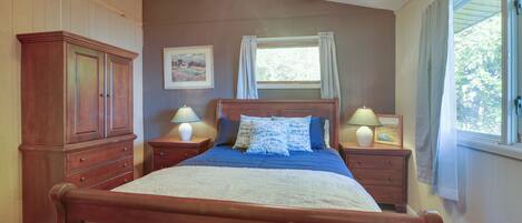 A bedroom with a wooden bed, two bedside tables with lamps, a window with curtains, and a wall-mounted picture.