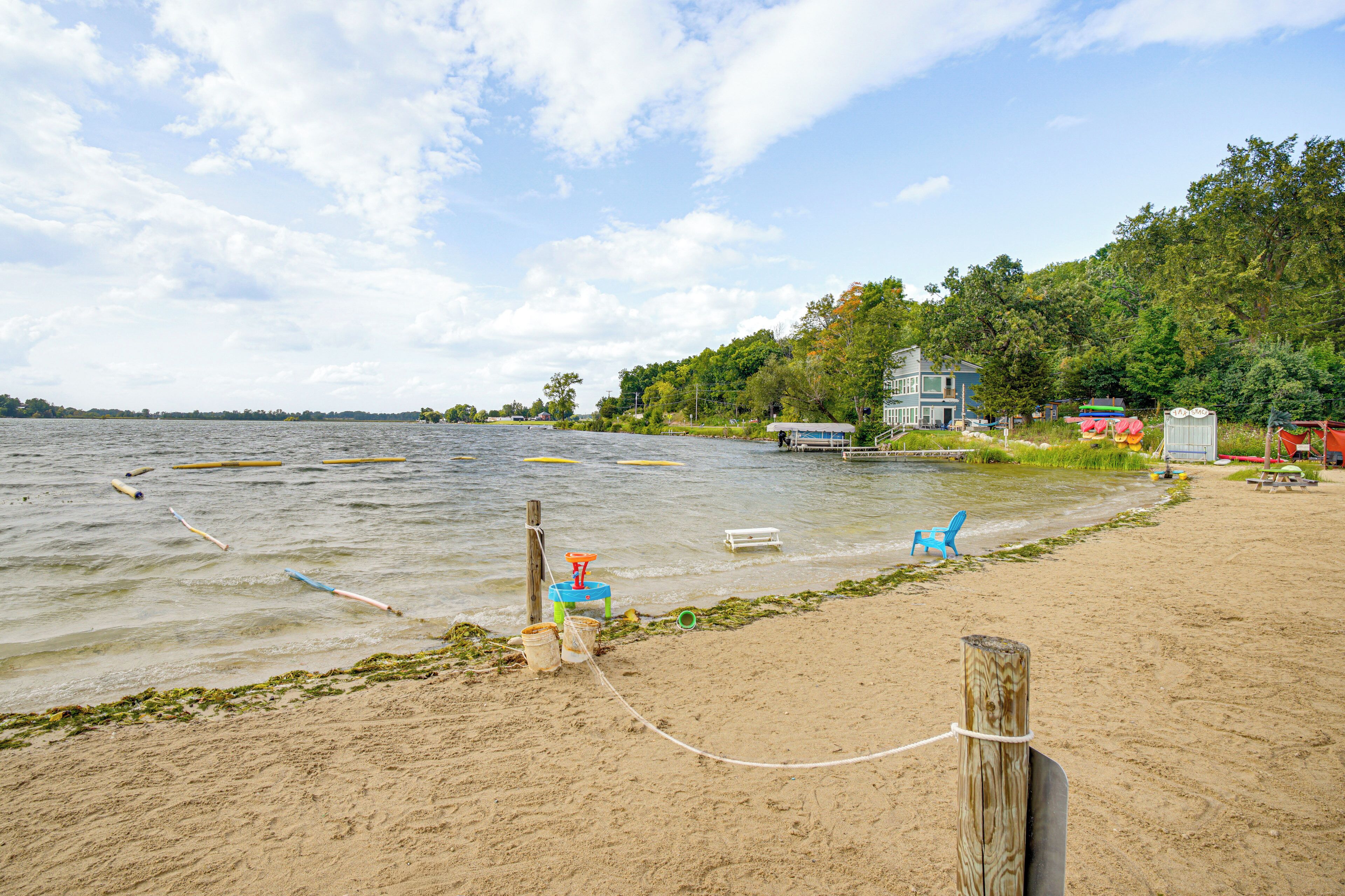 A sandy beach with a wooden post and a rope, a small dock with a blue chair, and a house with a red roof in the background.