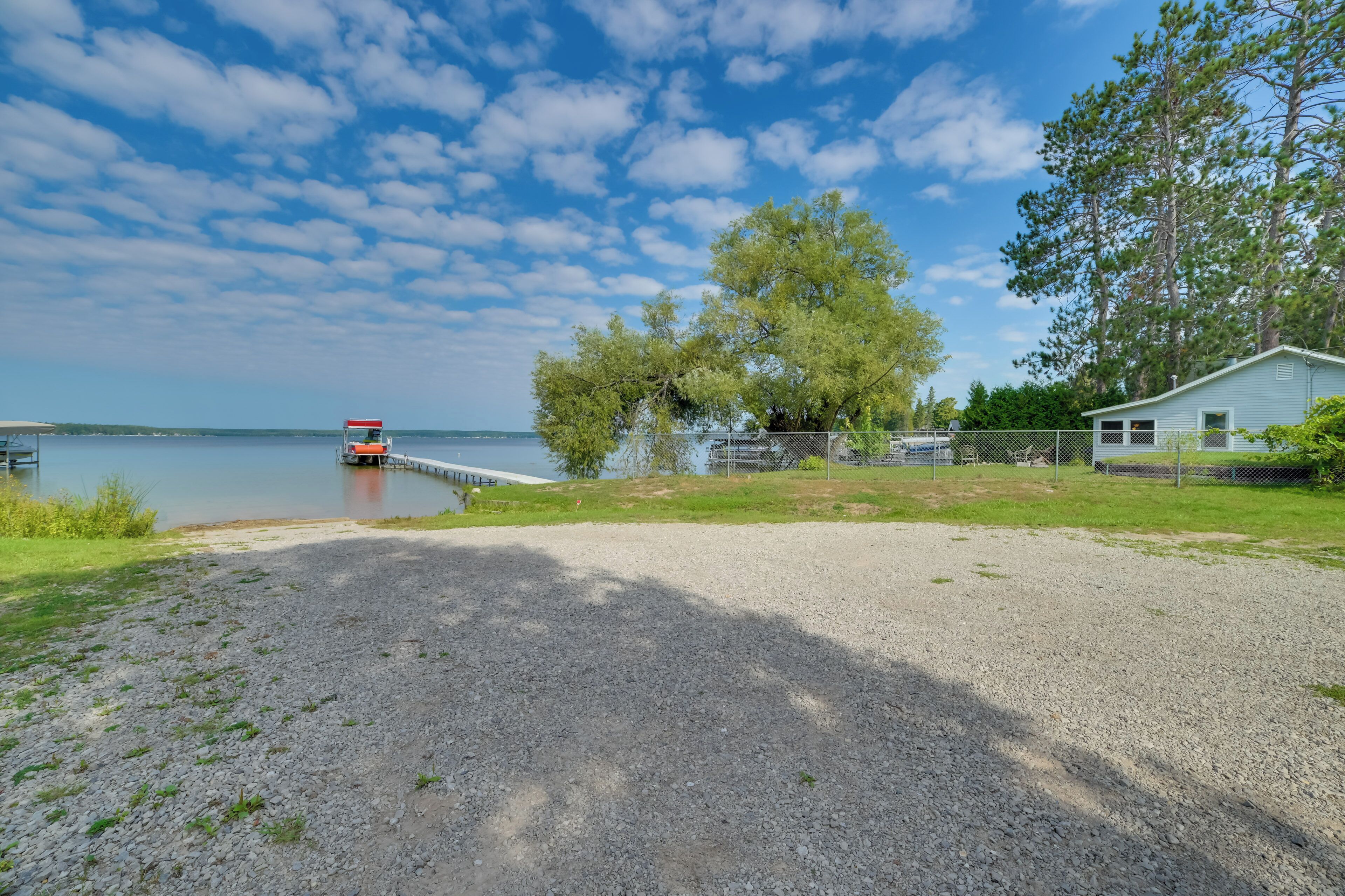 Une zone recouverte de gravier avec une jetée s’avançant dans un lac paisible, un petit bâtiment blanc et un grand arbre.