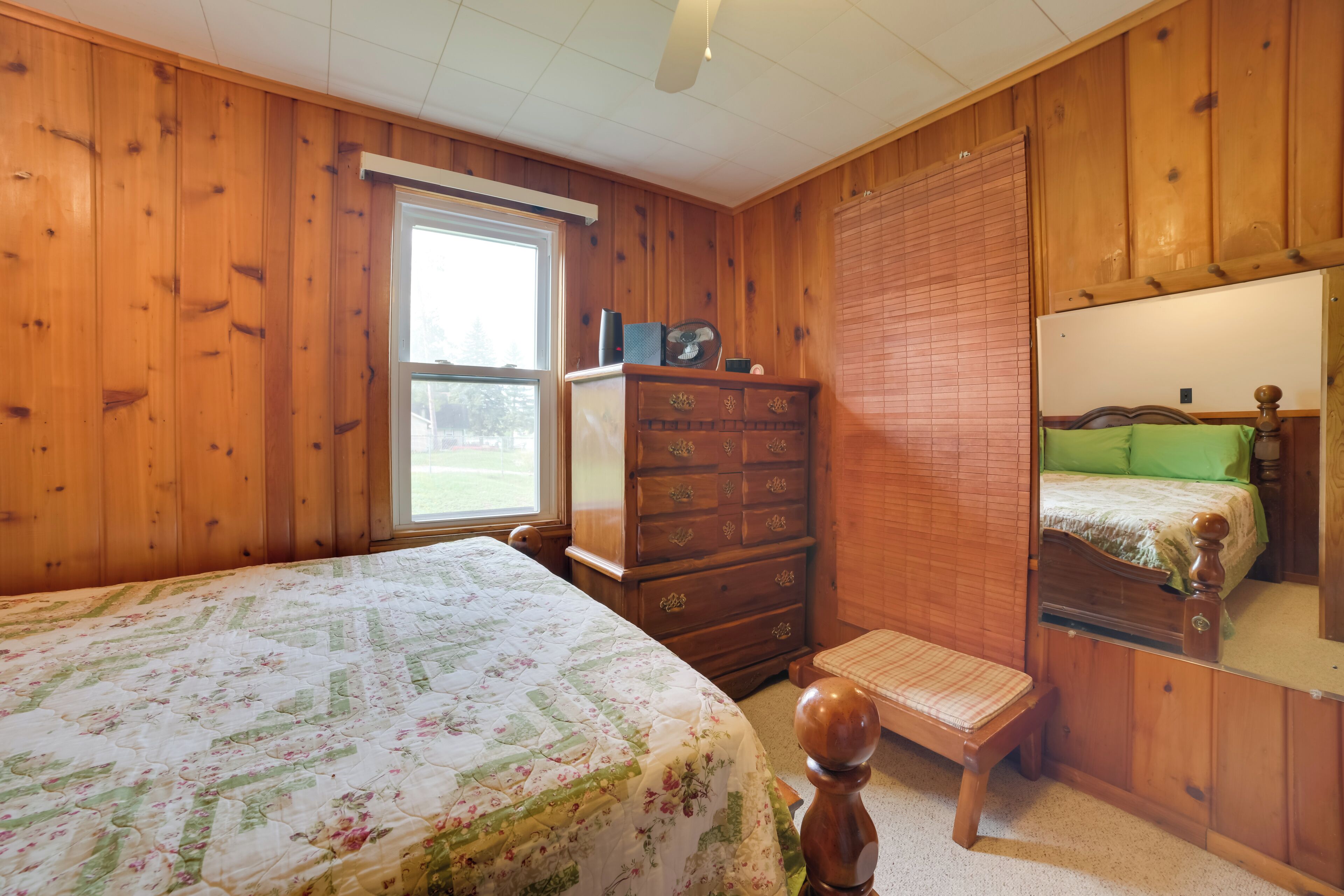 A bedroom with wooden paneling, a bed with a floral bedspread, a dresser, a mirror, and a window with blinds.