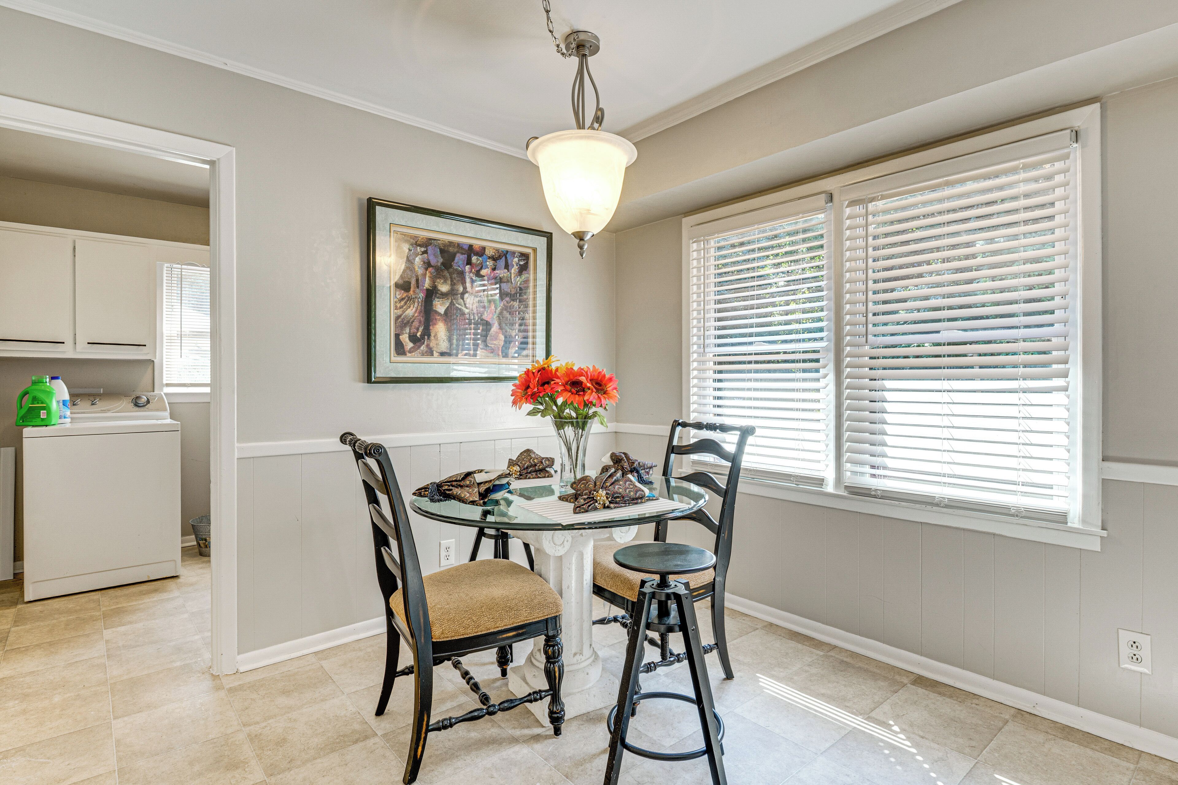 A dining area with a round glass table, four chairs, a vase with flowers, a painting on the wall, and a window with blinds.