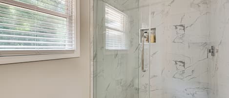 A glass-enclosed shower with marble walls, a window with blinds, and a vent on the wall.