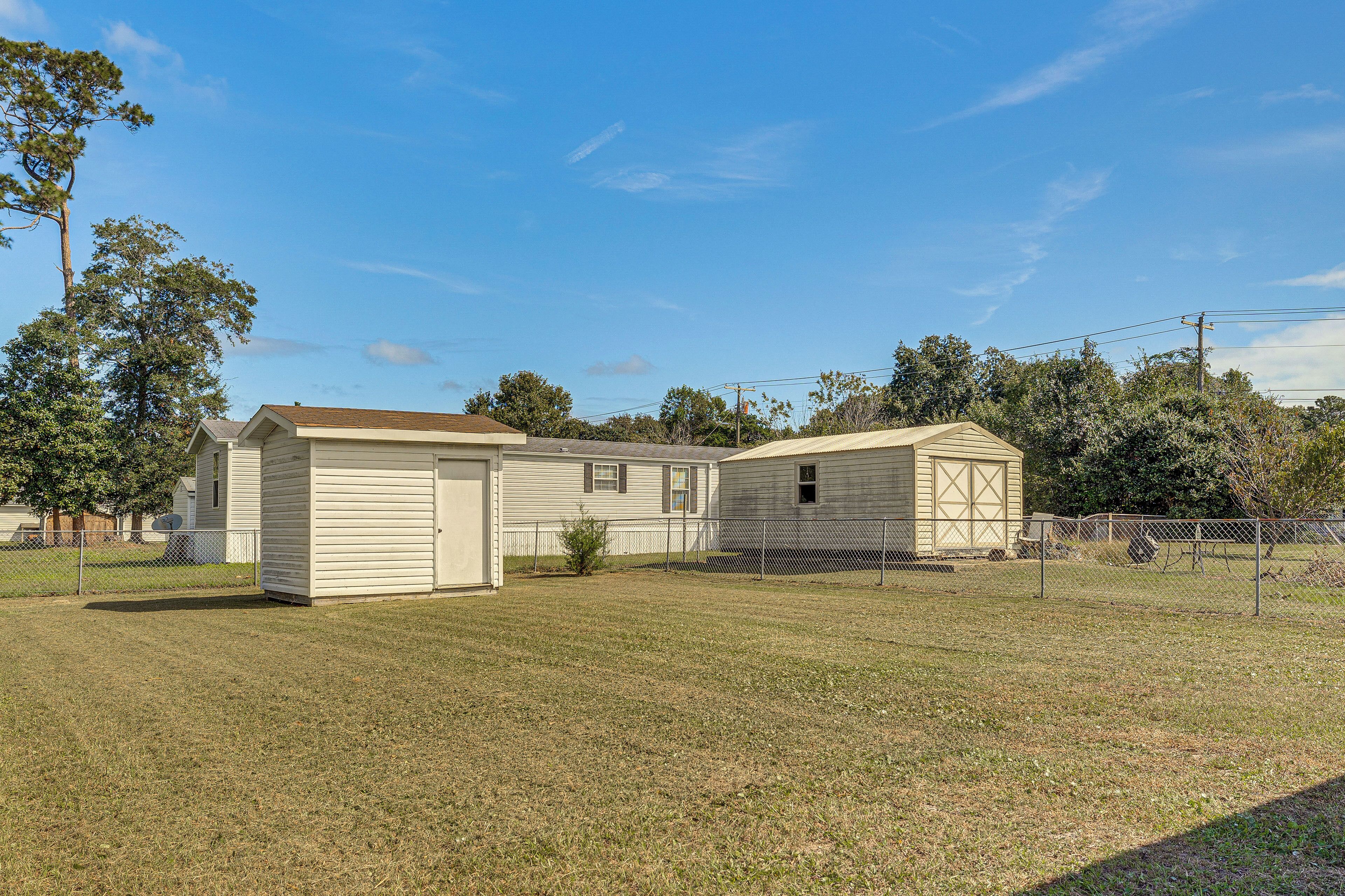 A single-story mobile home with a garage and a small attached shed, surrounded by a fenced yard and trees.