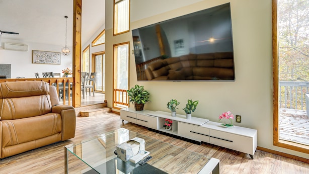 A modern living room with a large flat-screen TV mounted on the wall, a leather sofa, a glass coffee table, and a white entertainment unit with plants. - Luxury Home w/ Game Room, 4 Mi to Bushkill Falls! (Bushkill)