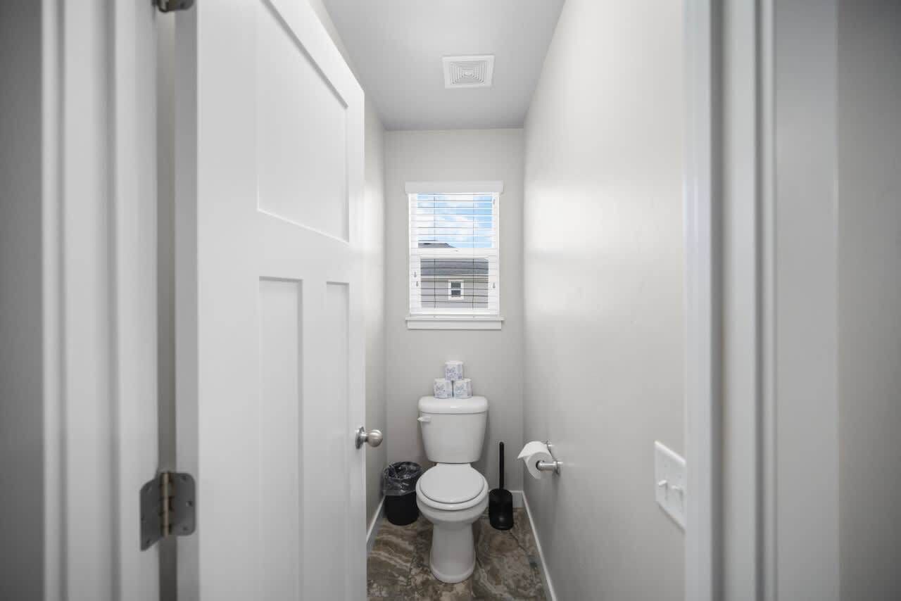 Ein schmaler Flur, der zu einem Badezimmer mit Toilette, Mülleimer und einem Fenster mit Jalousien führt.