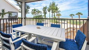Un área de estar al aire libre techada, con una mesa blanca y sillas azules, con vista a una playa con palmeras y un cielo despejado.