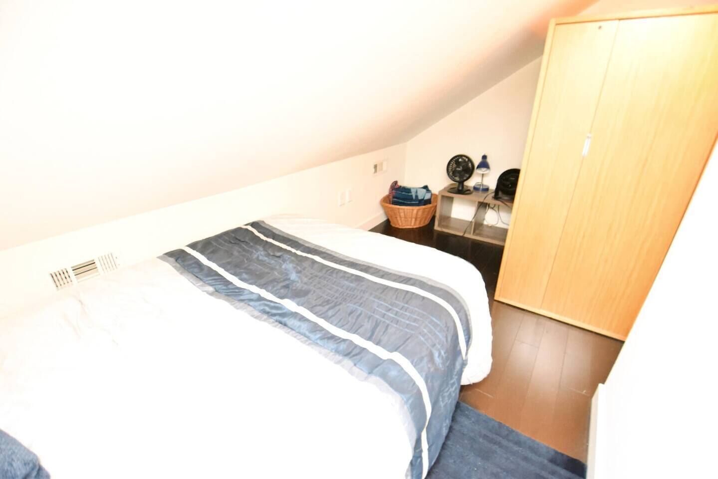 A bedroom with a bed, a wooden wardrobe, a small table with a lamp, and a wall clock.